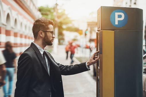 Young handsome businessman in glasses and the black business suit is paying his parking time using the automatic kiosk; the confident male employee is making payment with parking pay station terminal