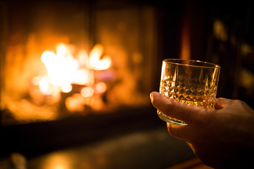Hand holding whiskey glass at warm fireplace