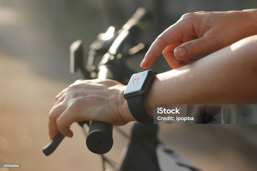 Woman cyclist hands with heart rate icon on smart watch, bicycle at summer trail. All on screen are design up. Cycling Stock Photo
