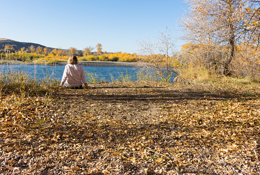 A woman seated on the bank looking at the Yellowstone River that is seen in the background. Fall foliage is on the distant riverbank.