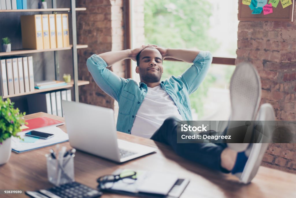 Jovem alegre afro freelancer está descansando num local de trabalho, com os pés em cima da mesa, com os olhos fechados, sorrindo, sonhando - Foto de stock de Escrivaninha royalty-free