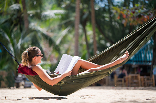 Relaxed woman sunbathing in hammock on the beach and reading a book.