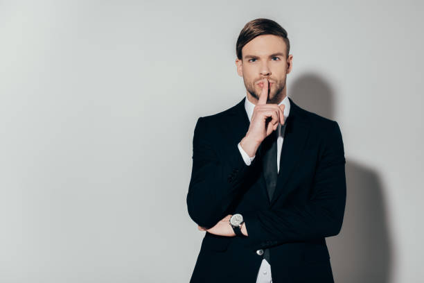 istock Young confident businessman in suit showing silence gesture on white background 928351502