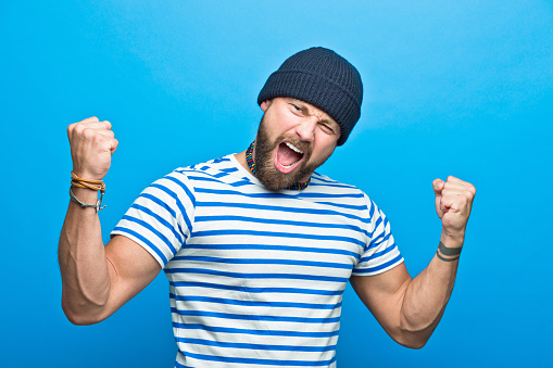 Portrait of happy bearded man wearing striped t-shirt and beanie hat flexing his muscles, screaming at camera. Studio shot, blue background.