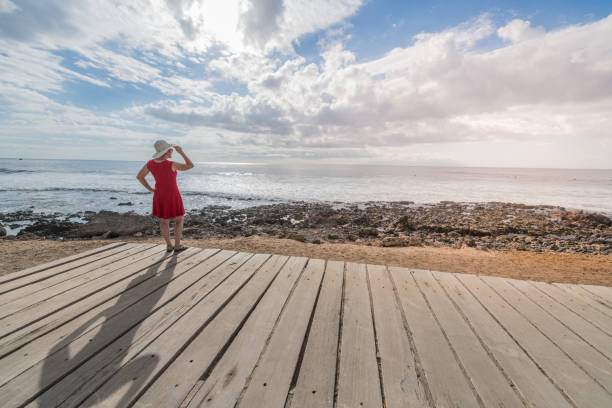 Senior woman with sun hat and red dress on the beach in Tenerife, Playa de las Americas, Spain. Europe. No logos. Nikon d850.