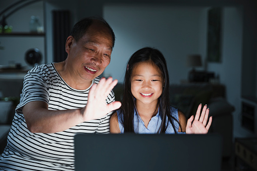 Little girl and her grandfather are using a laptop at home to video call their family. It is dark and the screen is illuminating their faces while they wave to the screen.