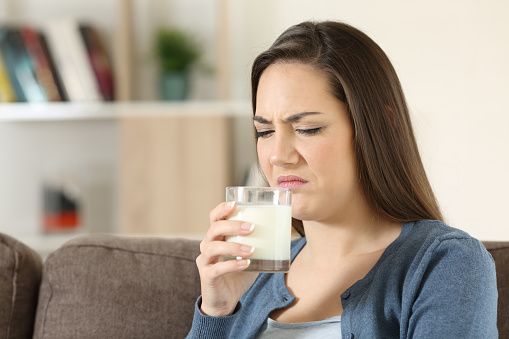 Disgusted woman tasing milk with bad flavor sitting on a couch in the living room at home