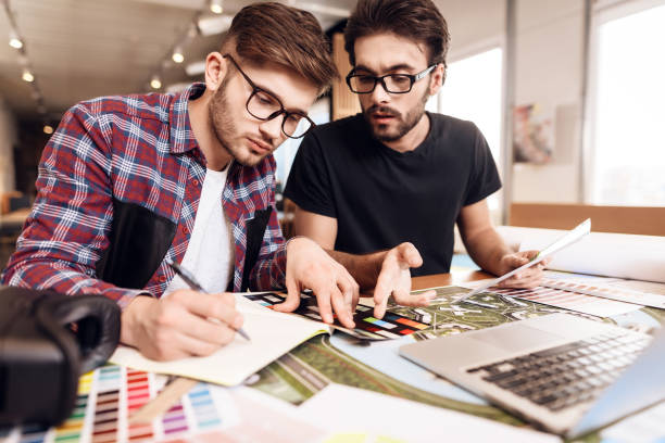 Two freelancer men looking at color swatches at laptop at desk. Two freelancer men in shirt and t-shirt looking at color swatches at laptop at desk. graphic designer photos stock pictures, royalty-free photos & images