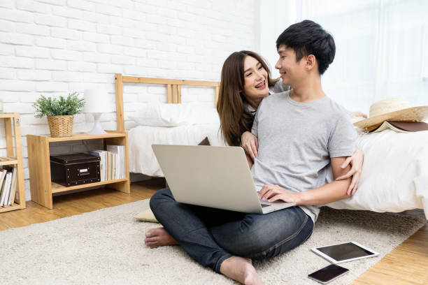 istock Young asian couple looking together using computer laptop internet shopping online or searching data honeymoon trip travel at bedroom in house.Happy lover hug each other and smiling on bed at  home 928458744