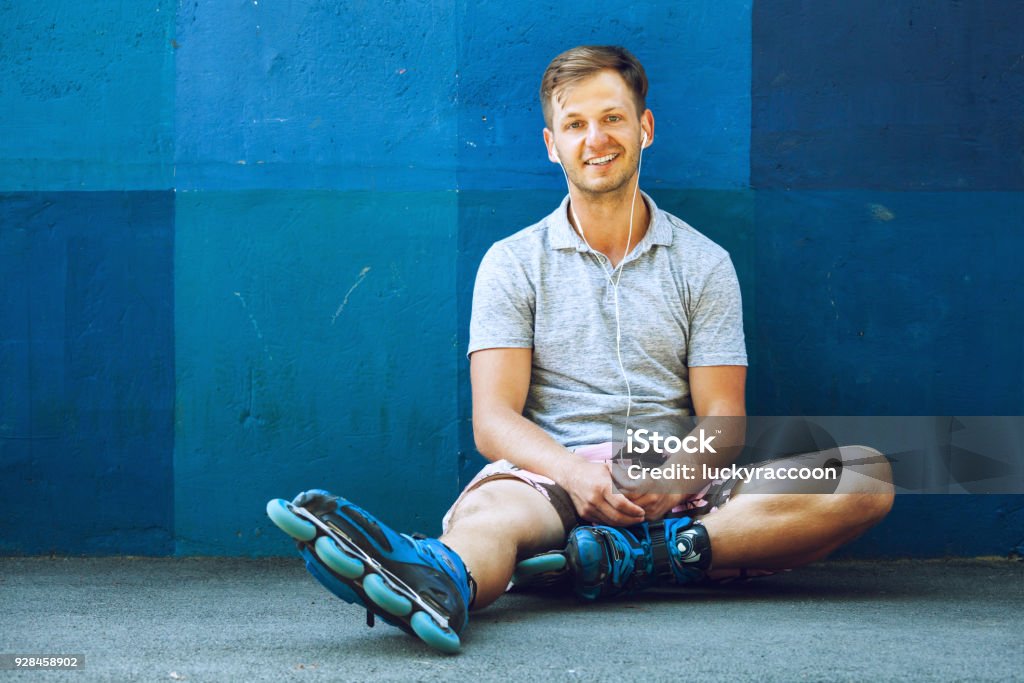 Happy handsome young roller in inline skates sitting against blue wall. Portrait of happy handsome young roller in inline skates sitting against blue wall. Inline Skate Stock Photo