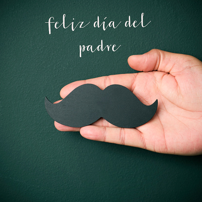 the text feliz dia del padre, happy fathers day in spanish, and the hand of a young caucasian man holding a mustache depicting a man face, on a dark green background