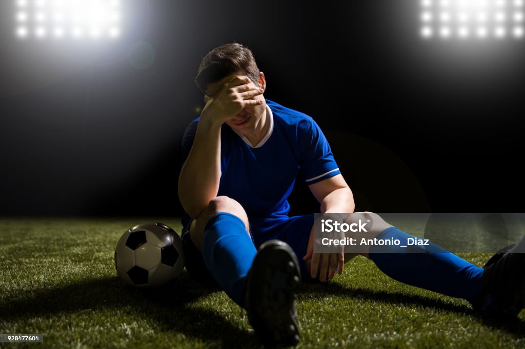 Footballer disappointed sitting on the grass field Disappointed soccer player in blue sitting on pitch after losing the match Soccer Stock Photo