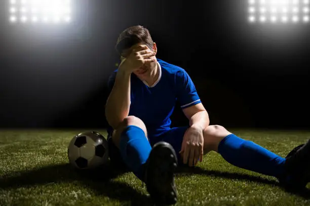Disappointed soccer player in blue sitting on pitch after losing the match