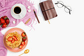 Beautiful flatlay arrangement with cup of coffee, hot waffles with cream and strawberries, glasses and other business accessories: concept of busy morning breakfast, white background.