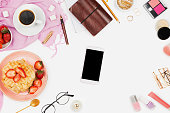 Beautiful flatlay arrangement with cup of coffee, hot waffles with cream and strawberries, smartphone with copyspace and beauty accessories: concept of busy morning breakfast, white background.