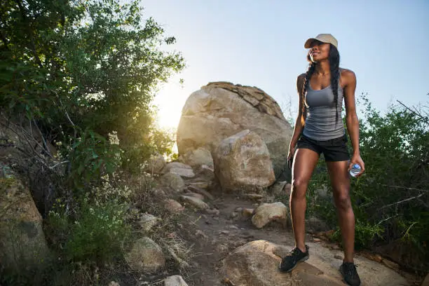 fit female hiker walking down rocky path while holding smartphone and waterbottle with sun behind her