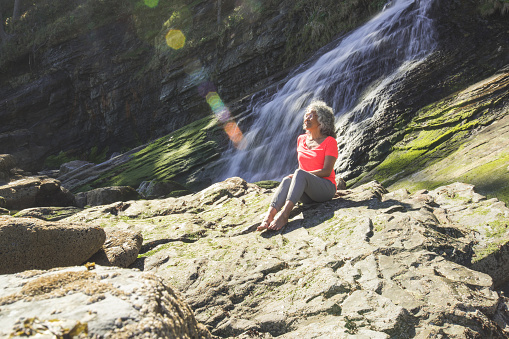 A cute ethnic senior woman relaxes by the ocean on the rocks by a waterfall. She has been hiking in the forest above the cliffs. The sunlight is breaking through the trees.