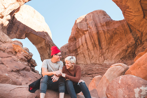 A young couple get up at sunrise to enjoy the majestic view at Arches National Park. They are sitting on a boulder and snuggled up and laughing together.  They are surrounded by natural rock formations and a rock bridge in the background.