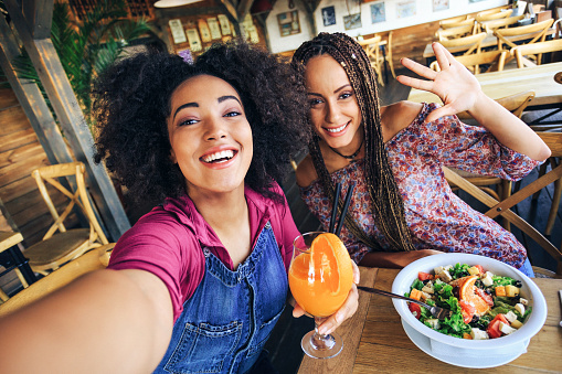 Young women in restaurant drinking juice and eating salad, taking selfie.
