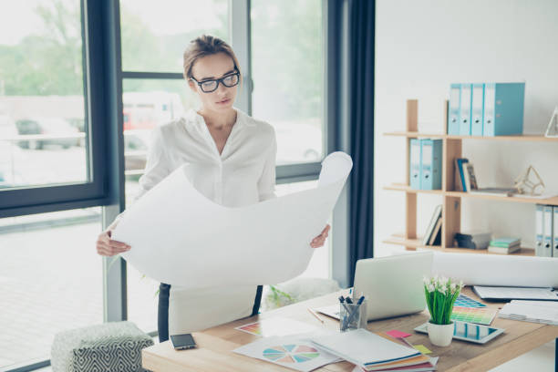 Creative brown haired serious focused concentrated lady is doing paper work, nice interior, huge windows, shelves with folders, perfect work station. Remodeling, visual art design, tone, web