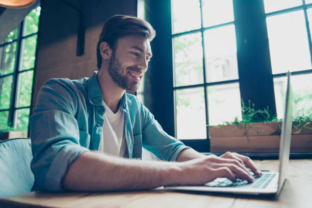 Side profile low angle shot of cheerful man smiling, searching information on his device in nice modern work station, in casual smart wear, grin, big windows with green nature view