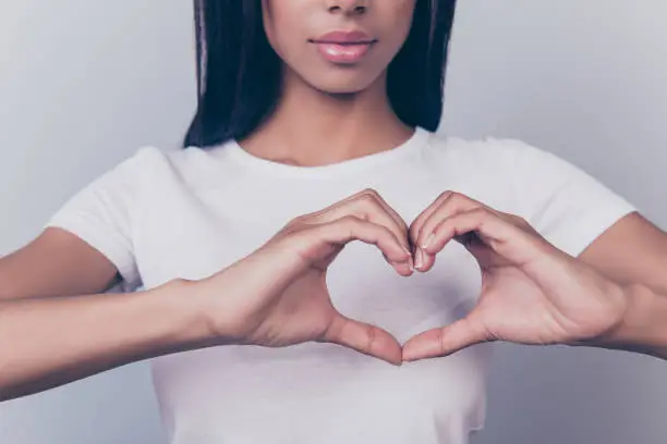 Cropped shot of cute brunette standing and showing heart gesture on the pure light grey background isolated, wearing casual white outfit, dark hair, pink full lips with nice pomade, french manicure