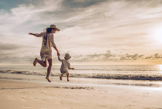 vista posterior de la feliz madre e hijo cogidos de la mano saltando en la playa. - family beach vacations travel fotografías e imágenes de stock