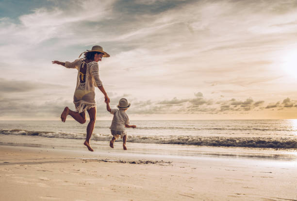 Rear view of happy mother and son holding hands while jumping on the beach. Back view of carefree mother and son holding hands while jumping on the beach at sunset. Copy space. beach play stock pictures, royalty-free photos & images