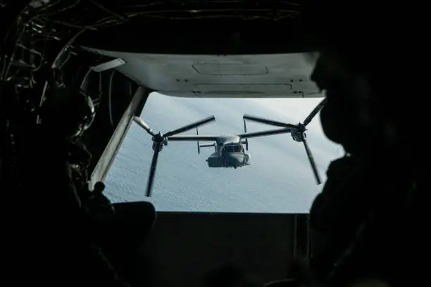 A US Marines V22 Osprey flying in close behind as we see out of the back of the one in front. Some Military personnel are silhouetted in the foreground.