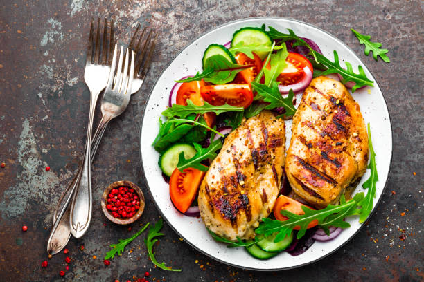 Photo of Grilled chicken breast. Fried chicken fillet and fresh vegetable salad of tomatoes, cucumbers and arugula leaves. Chicken meat with salad. Healthy food. Flat lay. Top view. Dark background