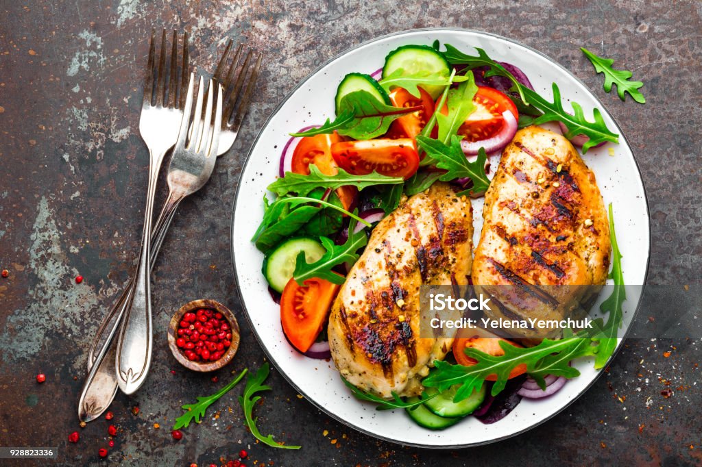 Grilled chicken breast. Fried chicken fillet and fresh vegetable salad of tomatoes, cucumbers and arugula leaves. Chicken meat with salad. Healthy food. Flat lay. Top view. Dark background Chicken Meat Stock Photo