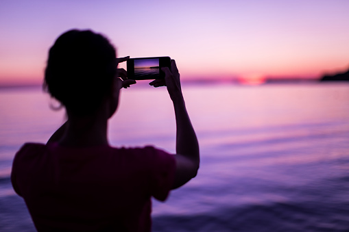 Rear View Of Pretty Young Woman Standing on Seaside and Taking Pictures Of Romantic Sunset