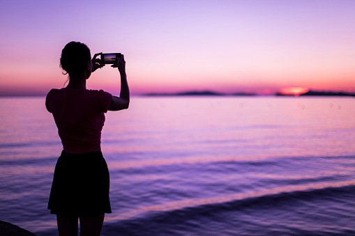 Rear View Of Pretty Young Woman Standing on Seaside and Taking Pictures Of Romantic Sunset,violet color photography