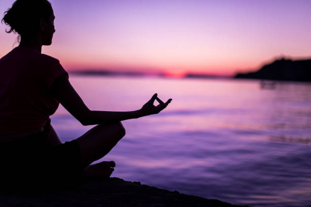 Young Woman Meditating By Peaceful Sea Calm Mindful Woman Meditating On Beach Faced Towards Majestic Sunset yoga lotus position meditating women stock pictures, royalty-free photos & images