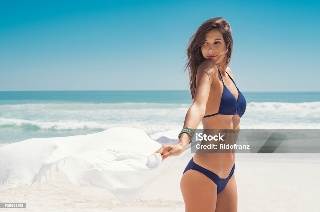 Woman enjoying breeze with scarf Young woman waving white scarf in wind at beach. Latin happy girl in blue bikini holding tissue and looking away at sea. Sexy fashion woman playing with the scarf on seashore. Women Stock Photo