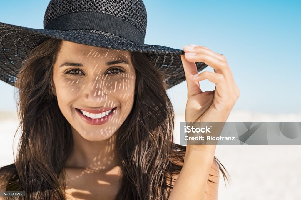 Woman wearing black hat at beach Portrait of beautiful young woman wearing summer black hat with large brim at beach. Closeup face of attractive girl with black straw hat. Happy latin woman smiling and looking at camera with sea in background. Women Stock Photo