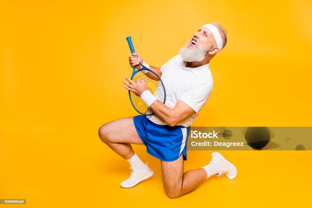 Sexy emotional cool pensioner grandpa practising rock music on a  sport equipment, stands on one knee, yell and shout. Body care, hobby, weight loss, lifestyle, strength and power, health Tennis Stock Photo