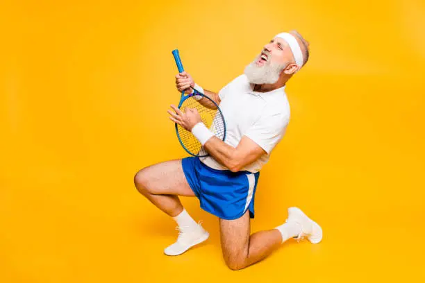 Photo of Sexy emotional cool pensioner grandpa practising rock music on a  sport equipment, stands on one knee, yell and shout. Body care, hobby, weight loss, lifestyle, strength and power, health