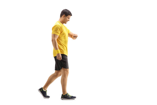 Full length profile shot of a young man walking and looking at his watch isolated on white background
