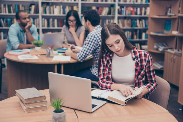 istock Focused concentrated attractive clever female student is learning with thesaurus in hard bookcase, wearing casual checkered shirt, behind her are classmates, book shelves of campus library 928932658