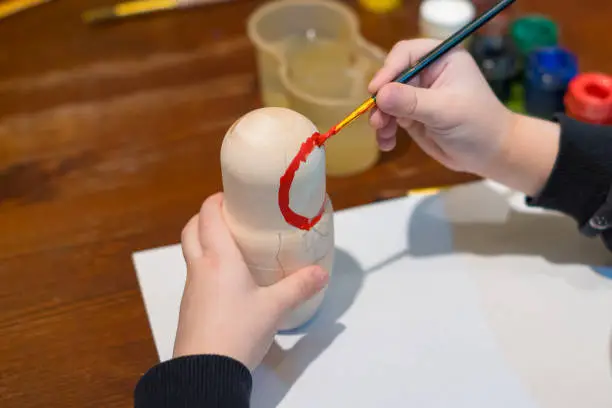 Photo of little girl paints russian nesting doll called matrioshka with red paint