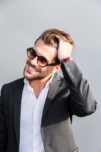 Male young handsome positive smiling businessman or worker in black suit with shirt in sunglasses with beard poses and adjusts corrects long hair with his hand outside in sunny day.