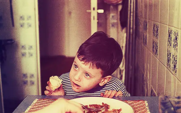 vintage color image of a cute boy eating in the kitchen and looking au at mommy.