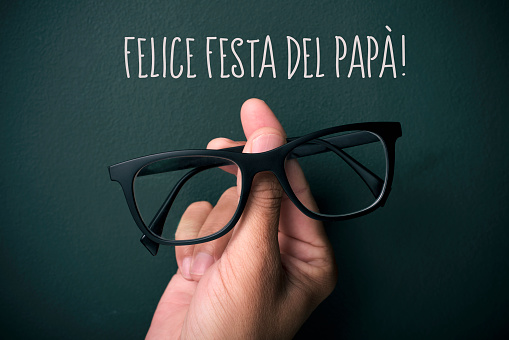 the text felice festa del papa, happy fathers day in italian, and the hand of a young caucasian man holding a pair of eyeglasses depicting a man face, on a dark green background