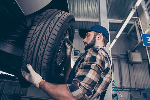 Low angle side profile shot of expert specialist technician changes tires, tyres of lifted up car, at auto workshop, wears checkered shirt, uniform costume, hat headwear