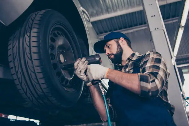 Photo of Technology, automotive, safety concept. Low angle shot of serious brunet bearded engineer in checkered shirt, hat head wear is torquing lugs nuts of wheel at a garage station, ceiling background