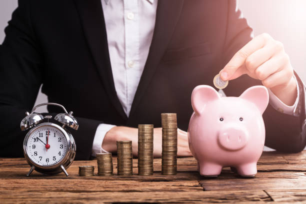 Businessperson's Hand Putting Coin In Piggybank A Businessperson's Hand Putting Coin In Piggybank Besides Alarm Clock On Desk race office stock pictures, royalty-free photos & images