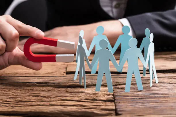 Businessperson's Hand Holding Red Horseshoe Magnet Attracting Paper Cut Out Figures On Wooden Desk