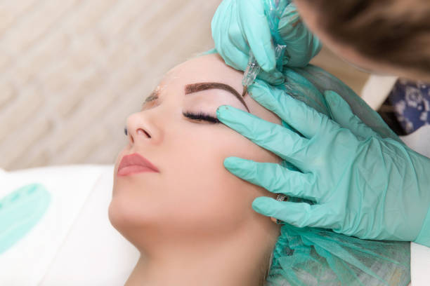 Microblading eyebrows work flow in a beauty salon. Woman having her eye brows tinted. Semi-permanent makeup for eyebrows. Focus on model's face and eyebrow stock photo
