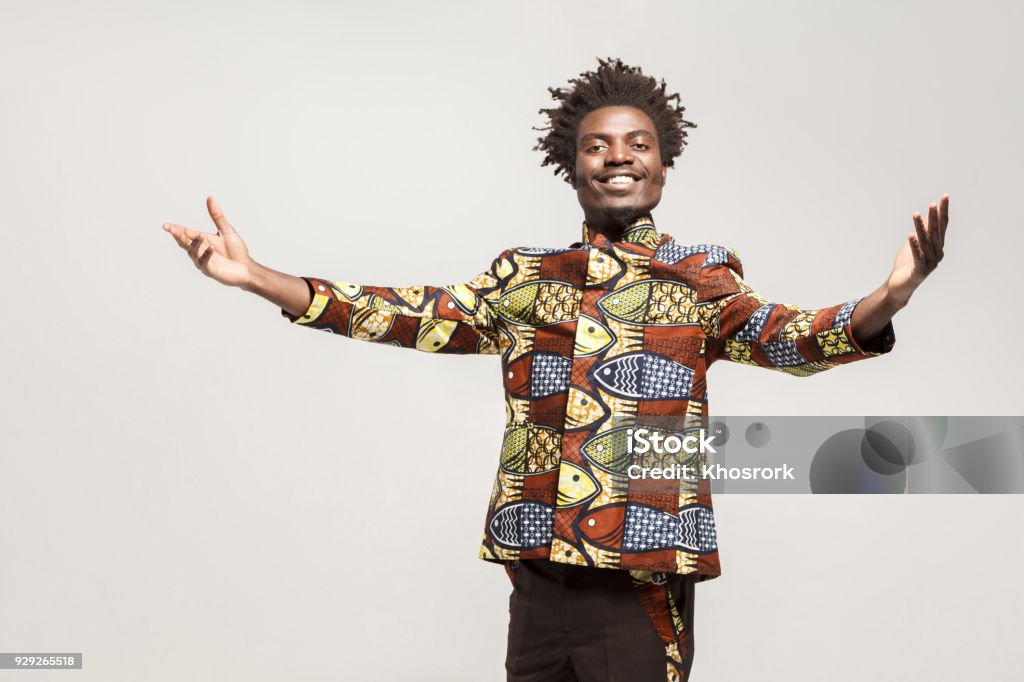 Friendly afro man in traditional clothes toothy smile, say welcome Friendly afro man in traditional clothes toothy smile, say welcome. Indoor, isolated on gray background Africa Stock Photo